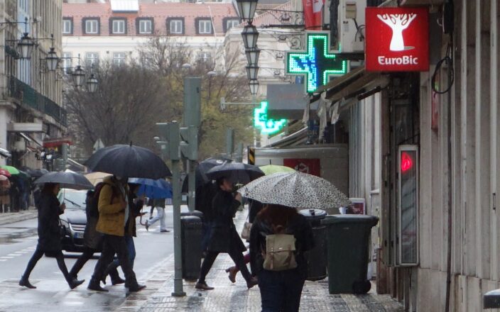 Pedestrians carry umbrellas across a rain-soaked Lisbon street beside shopfronts, glowing pharmacy crosses and traditional street lamps.