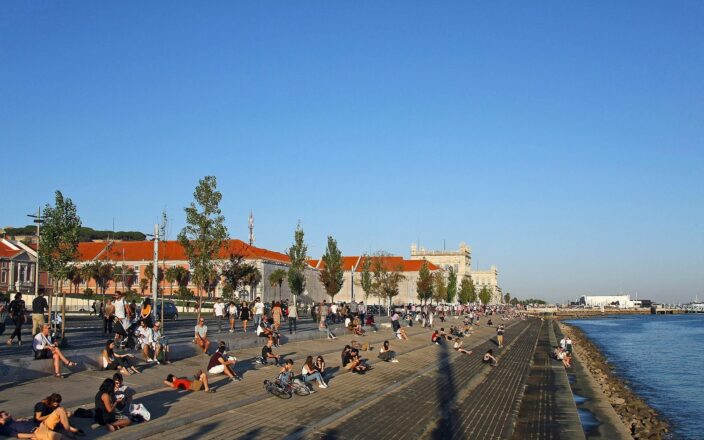 People stroll along Lisbon’s Ribeira das Naus promenade above broad riverside steps, with red-roofed buildings behind and the Tagus alongside.