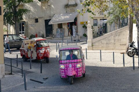 Pink and red tuk-tuks travel through a cobbled Alfama junction in Lisbon beneath trees near a São Jorge Castle sign.