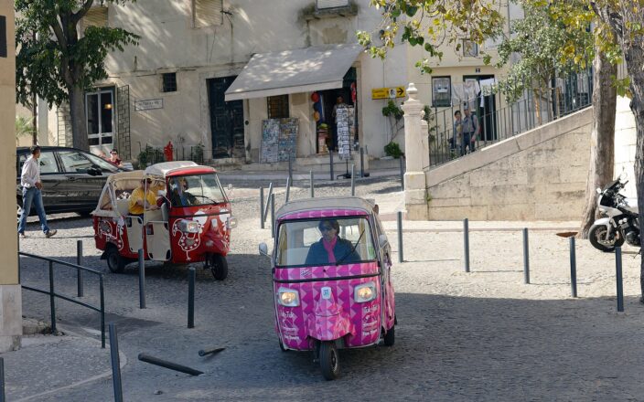Pink and red tuk-tuks travel through a cobbled Alfama junction in Lisbon beneath trees near a São Jorge Castle sign.
