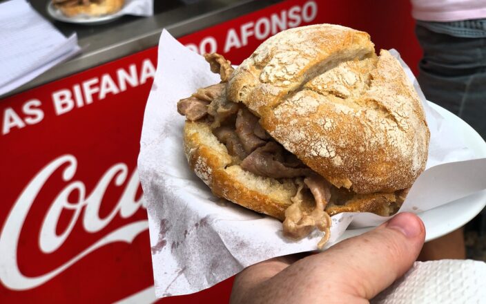 A pork-filled bifana in a crusty roll is held beside the As Bifanas do Afonso counter sign in Lisbon.