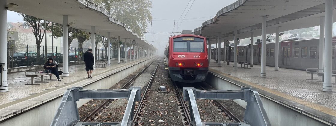 Red CP suburban train at Sintra station between covered platforms, with passengers waiting beside the tracks on a misty morning.