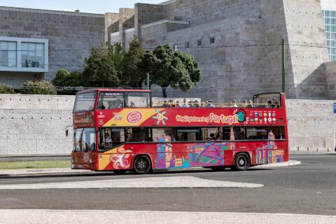 A red open-top sightseeing bus carries passengers past the pale stone walls of Centro Cultural de Belém in Lisbon.