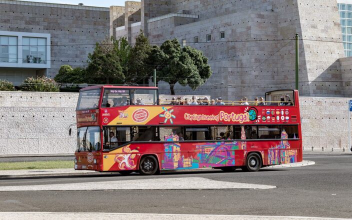 A red open-top sightseeing bus carries passengers past the pale stone walls of Centro Cultural de Belém in Lisbon.