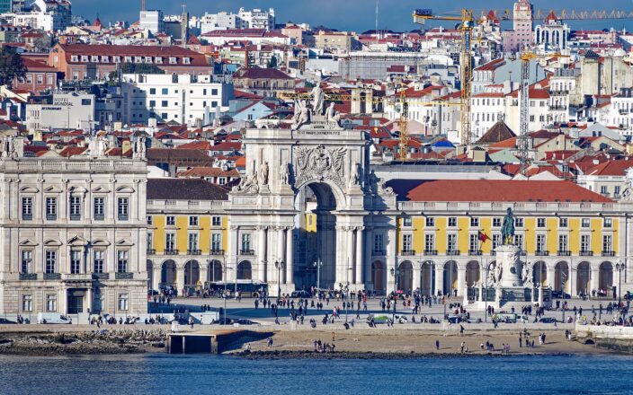 The Rua Augusta Arch and José I statue stand before yellow arcades at Lisbon’s Praça do Comércio, seen across the Tagus.