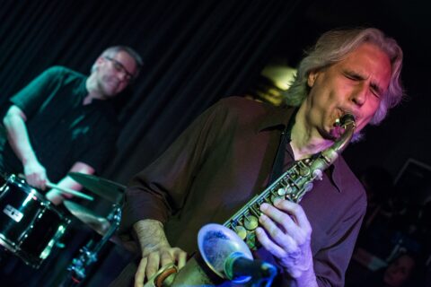 A saxophonist playing with closed eyes while a drummer plays behind him on a dark stage lit in purple and green at a Lisbon jazz club