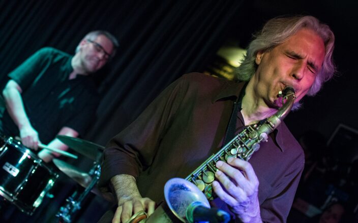 A saxophonist playing with closed eyes while a drummer plays behind him on a dark stage lit in purple and green at a Lisbon jazz club