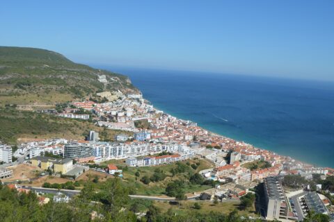 Sesimbra stretches between green hills and its blue bay, with white waterfront buildings, terracotta roofs and a boat offshore.