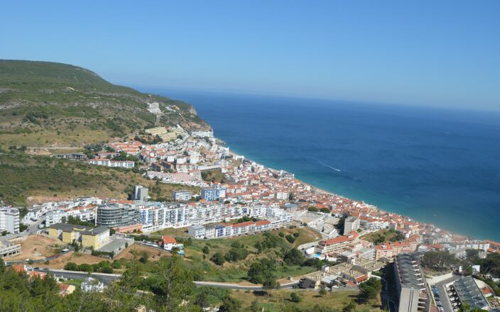 Sesimbra stretches between green hills and its blue bay, with white waterfront buildings, terracotta roofs and a boat offshore.