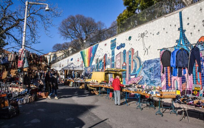 Shoppers browse records, bags and clothing beside the colorful tile mural at Feira da Ladra in Lisbon.