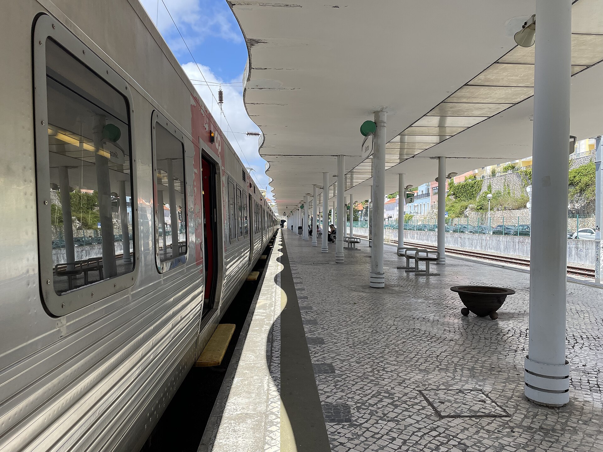 Silver suburban train beside Sintra station platform, with white canopy columns, patterned paving and passengers seated farther along.