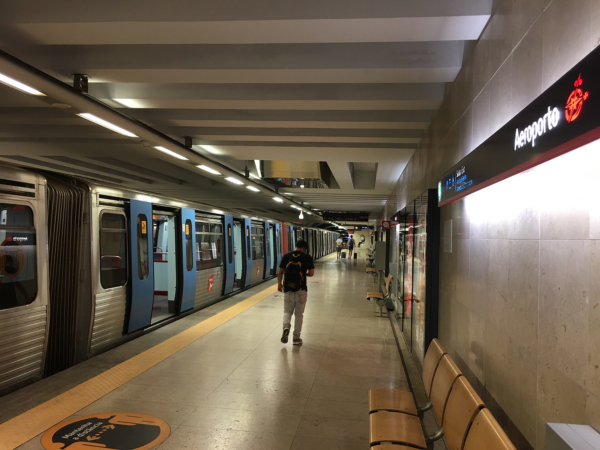 A silver train with blue doors waits at Lisbon Metro Aeroporto station while a passenger walks along the platform.