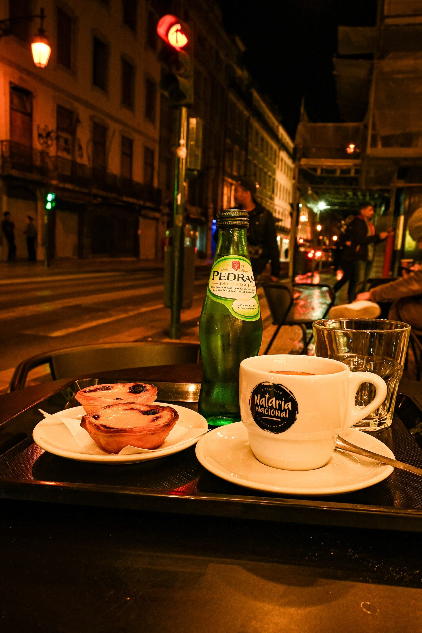 Small coffee cup and two pastéis de nata on a tray beside a bottle of Pedras mineral water at a pavement table on Rua Augusta in Lisbon at night.
