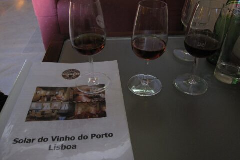 Three glasses of port stand beside a Solar do Vinho do Porto Lisboa booklet on a glass table in Lisbon.