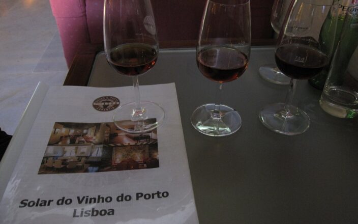 Three glasses of port stand beside a Solar do Vinho do Porto Lisboa booklet on a glass table in Lisbon.