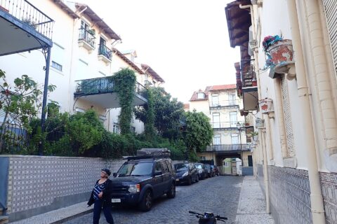Tiled walls and leafy iron balconies line the cobbled lane of Vila Berta in Lisbon, with a cyclist beside parked cars.
