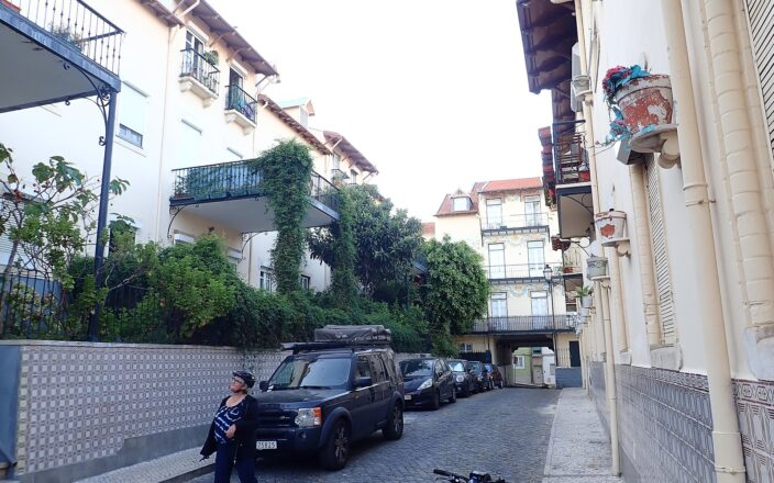Tiled walls and leafy iron balconies line the cobbled lane of Vila Berta in Lisbon, with a cyclist beside parked cars.
