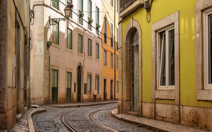 Tram rails curve along a narrow cobbled street between pastel houses and iron balconies in Alfama, Lisbon.