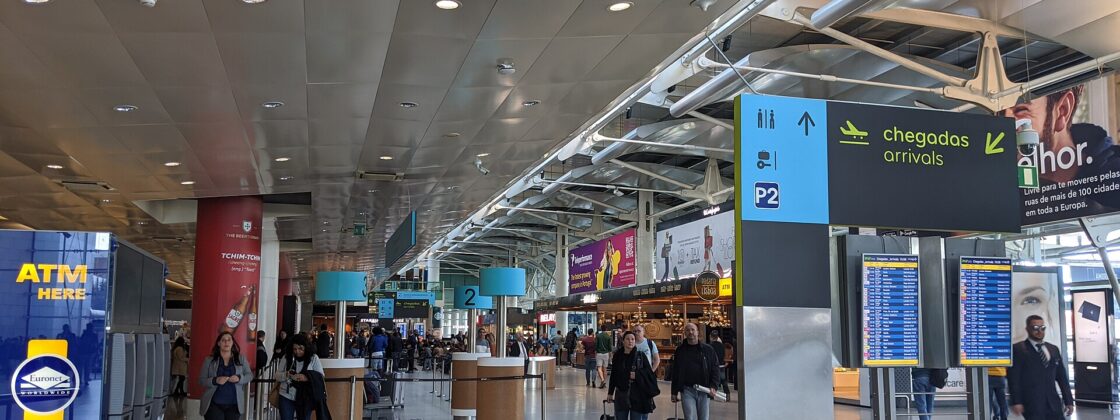 Travelers walk through Lisbon Airport beneath bilingual arrivals signs and flight information screens in the terminal hall.