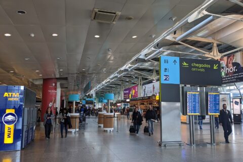 Travelers walk through Lisbon Airport beneath bilingual arrivals signs and flight information screens in the terminal hall.