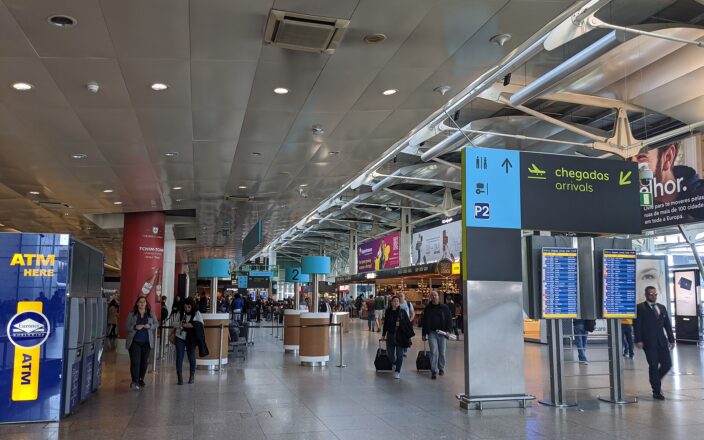 Travelers walk through Lisbon Airport beneath bilingual arrivals signs and flight information screens in the terminal hall.