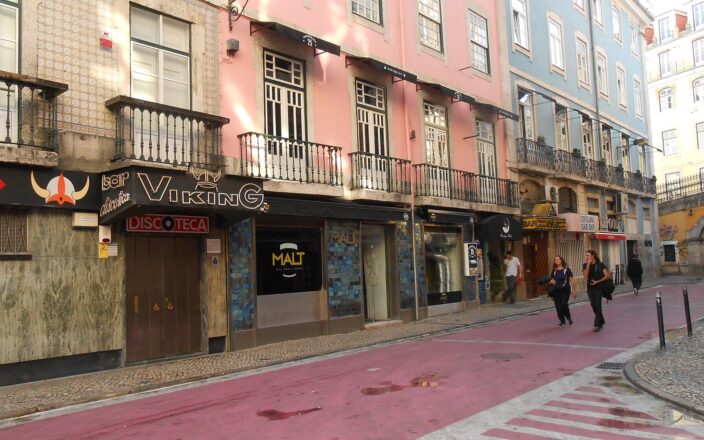 Viking disco and neighboring bars line the pink-painted Rua Nova do Carvalho in Lisbon, with pedestrians passing beneath pastel facades.