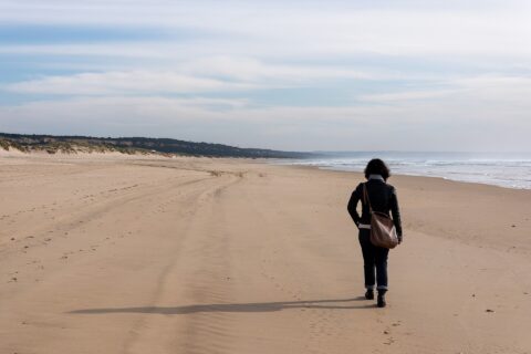 A visitor walks along Rainha Beach on Costa da Caparica, with Atlantic waves, low dunes and wooded cliffs in the distance.
