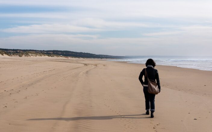 A visitor walks along Rainha Beach on Costa da Caparica, with Atlantic waves, low dunes and wooded cliffs in the distance.