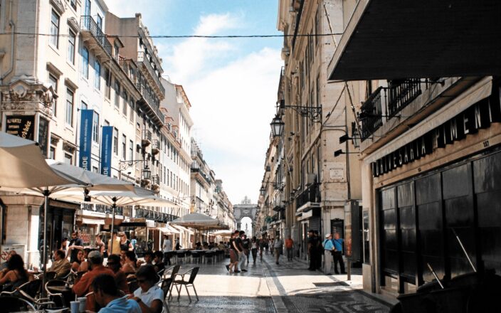 Visitors and cafe tables line Lisbon’s Rua Augusta, with patterned paving leading toward the triumphal arch.