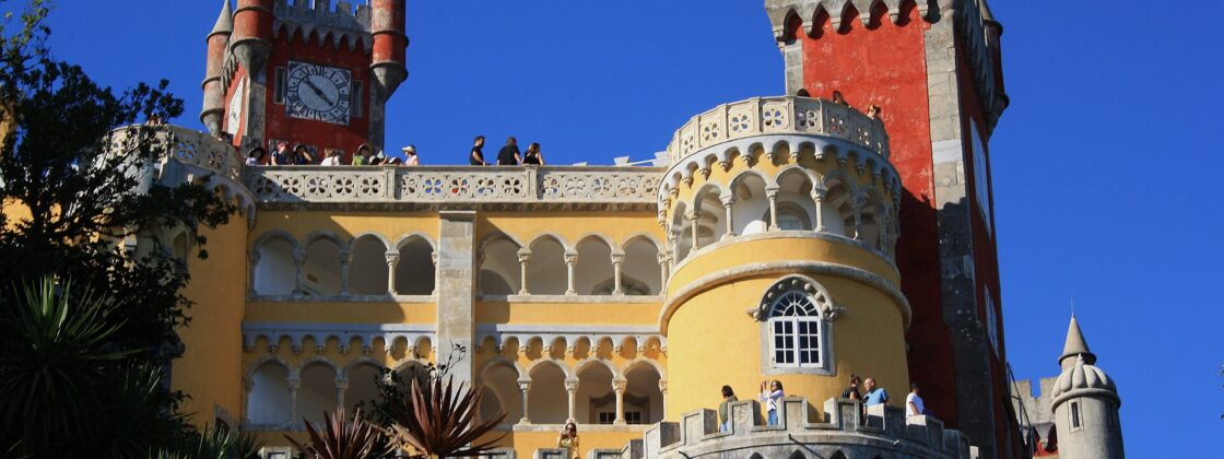 Visitors explore Pena Palace’s sunny terraces in Sintra, framed by yellow arcades, red towers and ornate stone balustrades.