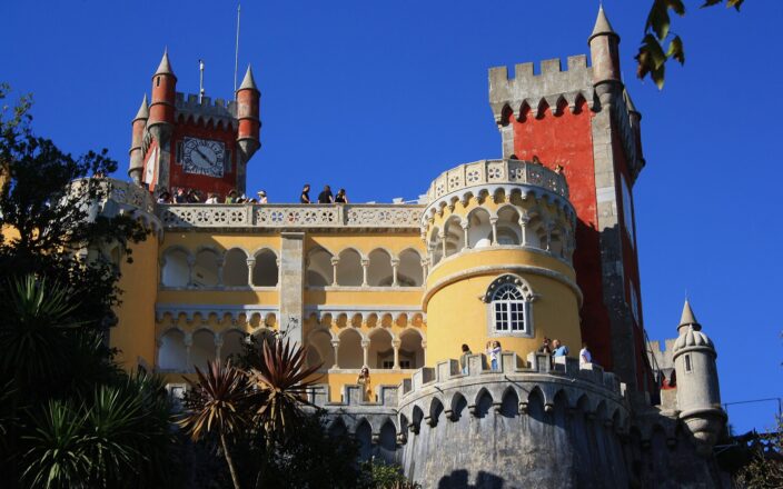 Visitors explore Pena Palace’s sunny terraces in Sintra, framed by yellow arcades, red towers and ornate stone balustrades.