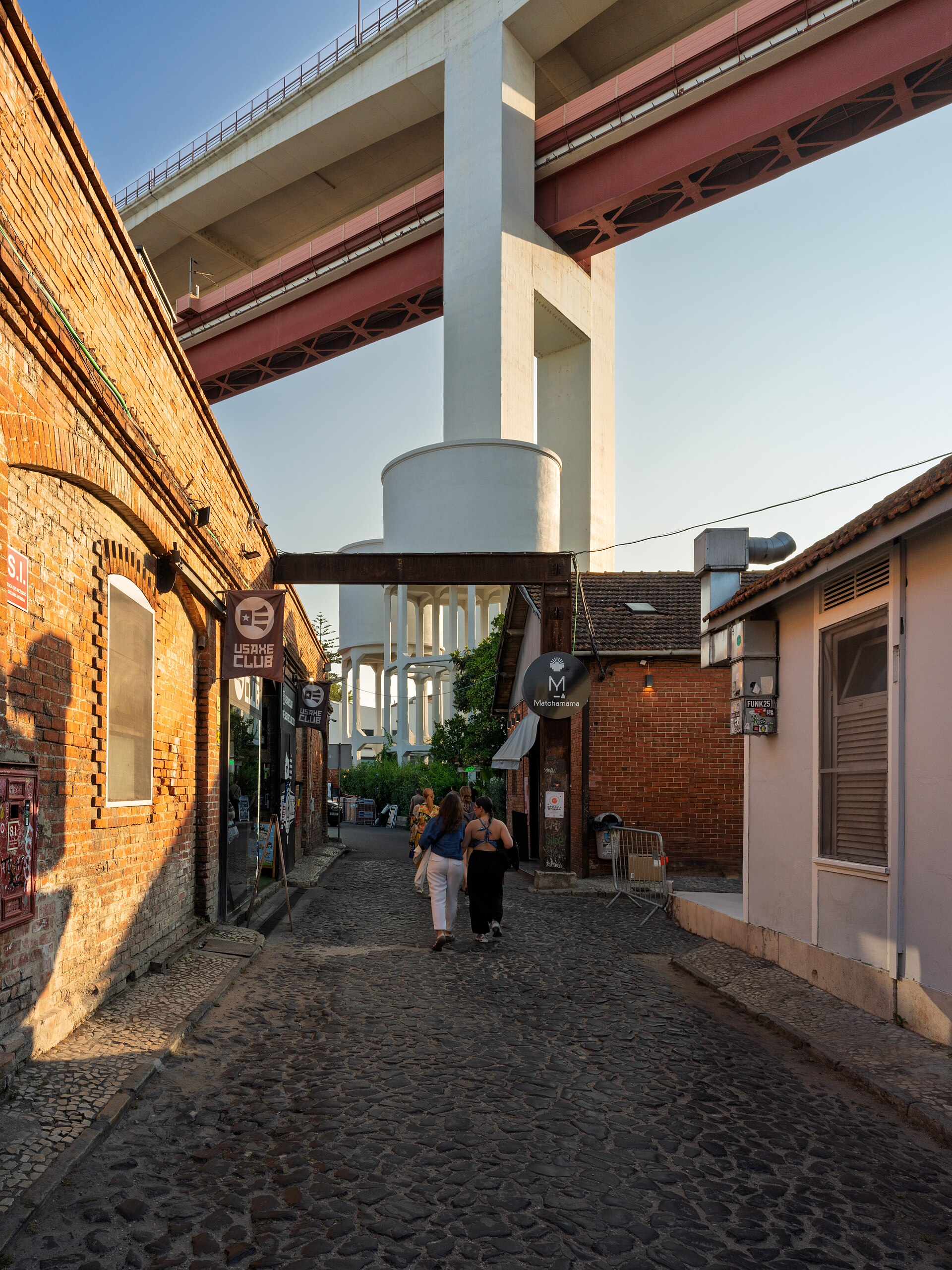Visitors follow a brick-lined alley at LX Factory in Lisbon beneath the 25 de Abril Bridge approach and beside its water tower.