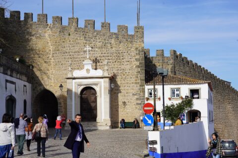 Visitors gather outside the Porta da Vila in Óbidos, beneath crenellated stone walls beside whitewashed houses with blue trim.