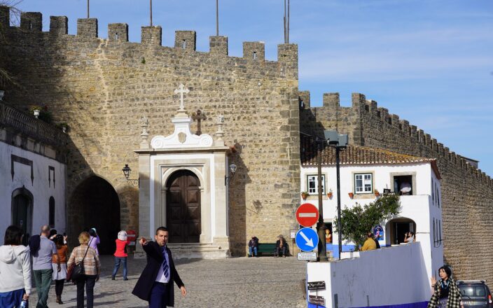 Visitors gather outside the Porta da Vila in Óbidos, beneath crenellated stone walls beside whitewashed houses with blue trim.