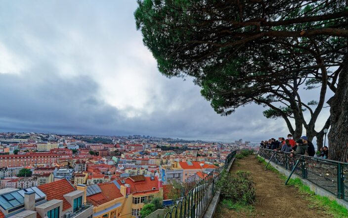 Visitors look across Lisbon rooftops from the pine-shaded terrace at Miradouro da Senhora do Monte beneath cloudy skies.