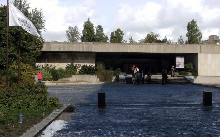 Visitors outside Lisbon's Calouste Gulbenkian Museum, where a low concrete entrance overlooks a planted pool and cobbled forecourt.