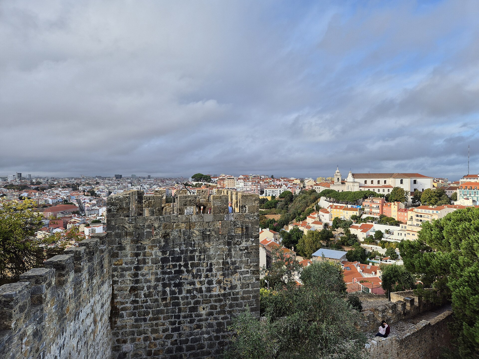 Visitors stand behind São Jorge Castle's battlements above Lisbon's red rooftops, with a white church on the neighboring hillside.