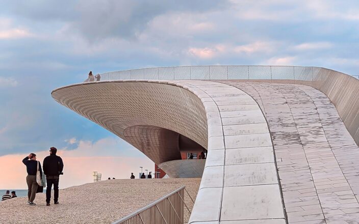 Visitors stand beside MAAT’s sweeping ceramic-clad roof in Lisbon, with more people visible along the curved rooftop railing.