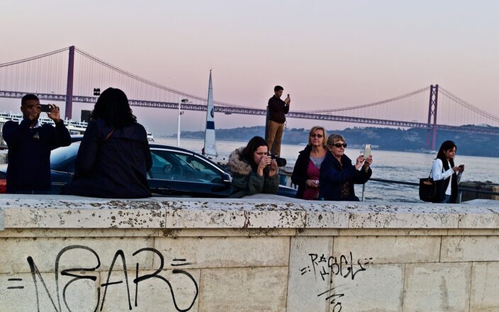 Visitors use smartphones and a camera on Lisbon’s waterfront at dusk, with the 25 de Abril Bridge spanning the Tagus behind them.