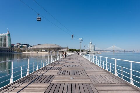 Visitors walk along Lisbon’s Parque das Nações boardwalk beneath cable cars, with the Vasco da Gama Tower and Bridge ahead.