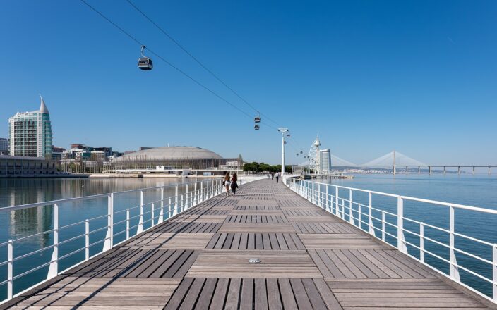 Visitors walk along Lisbon’s Parque das Nações boardwalk beneath cable cars, with the Vasco da Gama Tower and Bridge ahead.