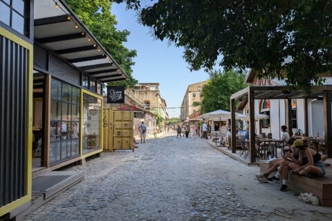 Visitors walk along LX Factory’s cobbled main street in Lisbon, between converted warehouses, shops and shaded café terraces.