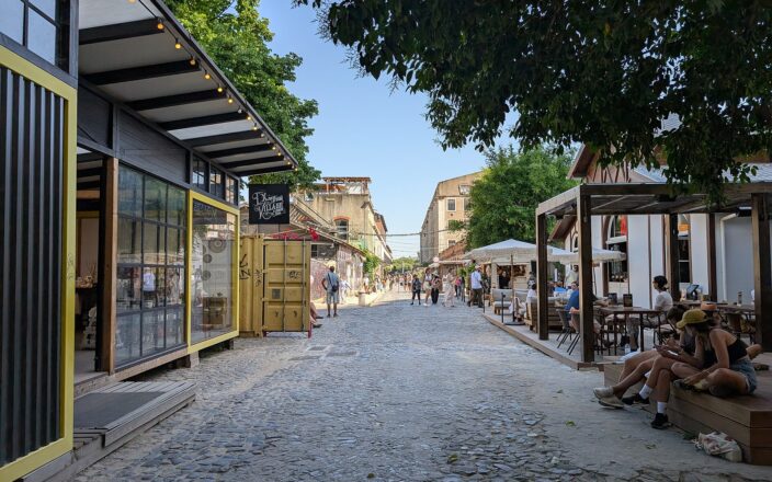 Visitors walk along LX Factory’s cobbled main street in Lisbon, between converted warehouses, shops and shaded café terraces.