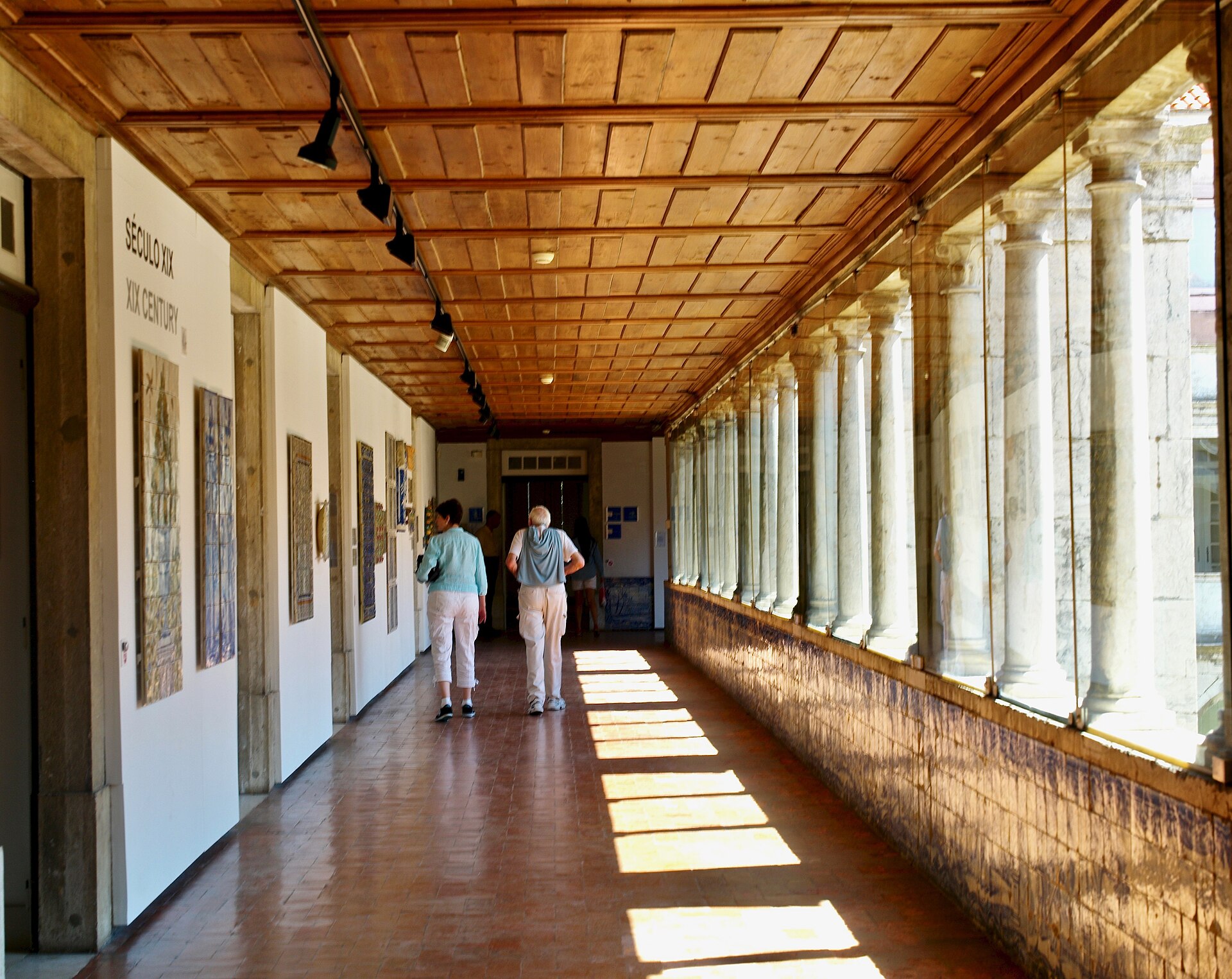 Visitors walk past azulejo panels in Lisbon's National Tile Museum, beneath a wooden ceiling beside a sunlit glazed cloister arcade.
