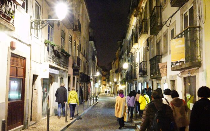 Walkers seen from behind on a narrow cobbled street in Bairro Alto at night, lit by wrought-iron wall lanterns between tall old townhouses.