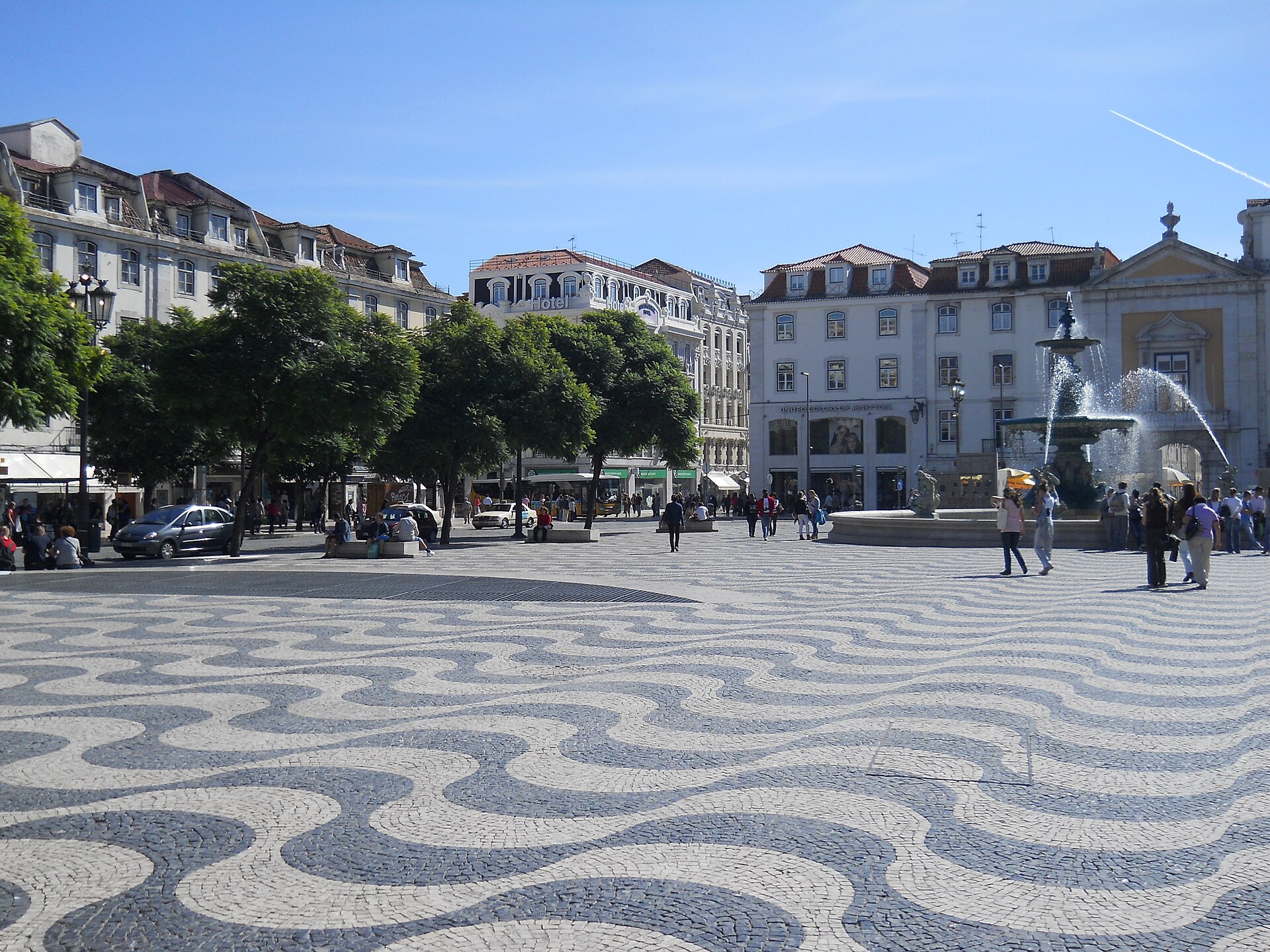 Wave patterned black and white paving fills Rossio Square in Lisbon, with pedestrians beside a spraying fountain and tree lined buildings.