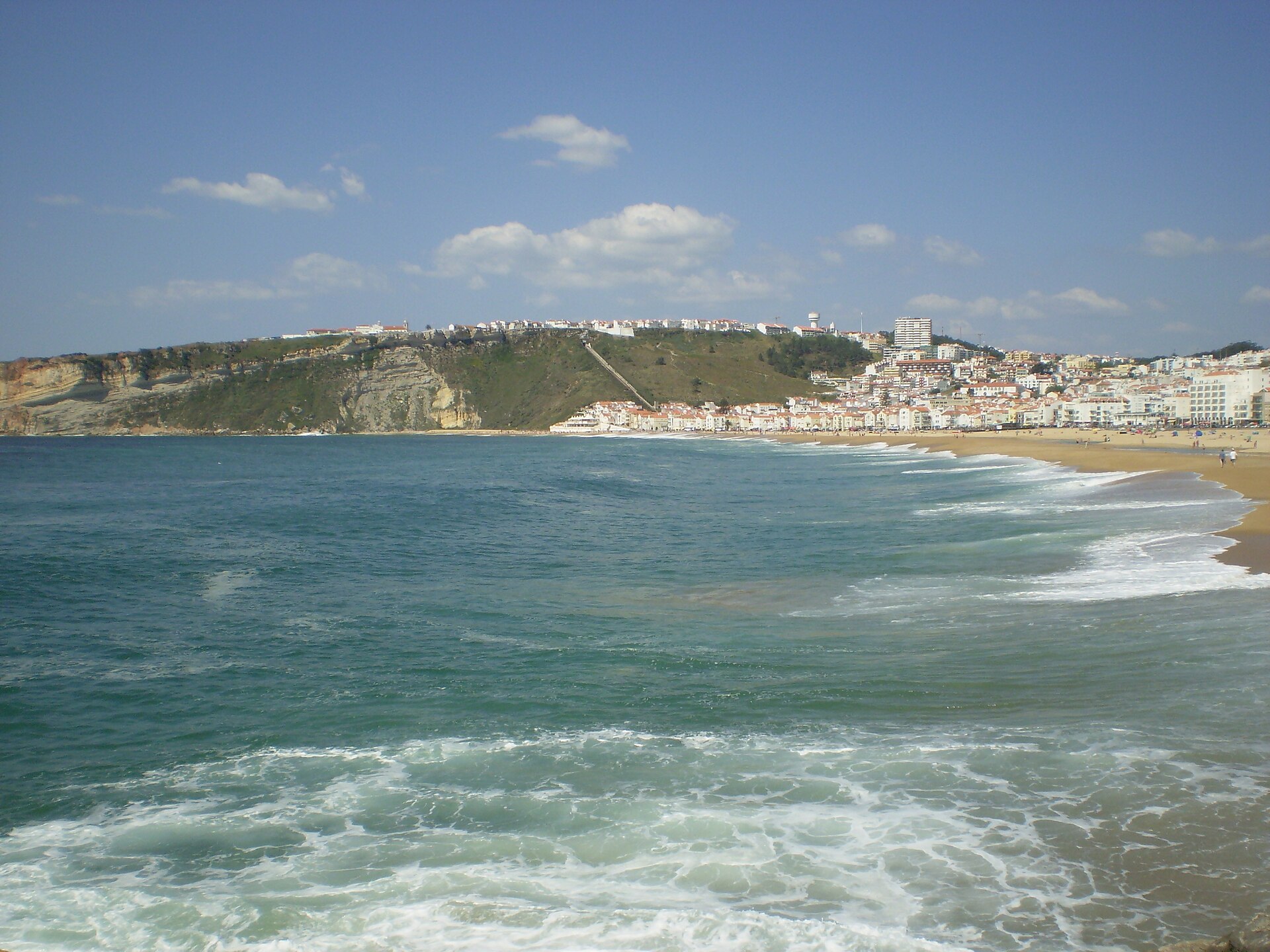Waves wash onto Nazaré’s sandy town beach beneath the Sítio headland, with the funicular track visible on the slope.