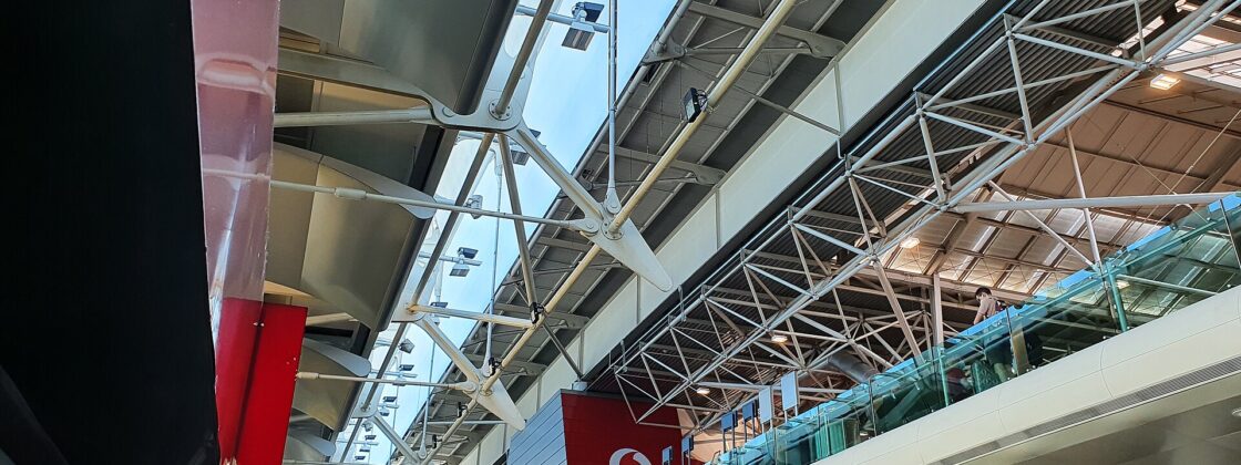 White roof trusses span Lisbon Airport Terminal 1 above signs pointing toward departures, Terminal 2 and ground transport.
