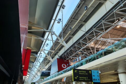 White roof trusses span Lisbon Airport Terminal 1 above signs pointing toward departures, Terminal 2 and ground transport.