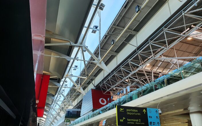 White roof trusses span Lisbon Airport Terminal 1 above signs pointing toward departures, Terminal 2 and ground transport.